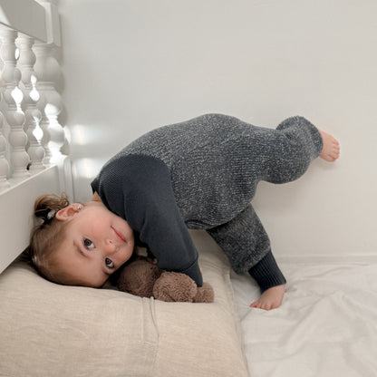 A toddler wearing a dark gray heathered knit one-piece jumpsuit crawls on a white bed, resting on a beige pillow and holding a brown teddy bear.