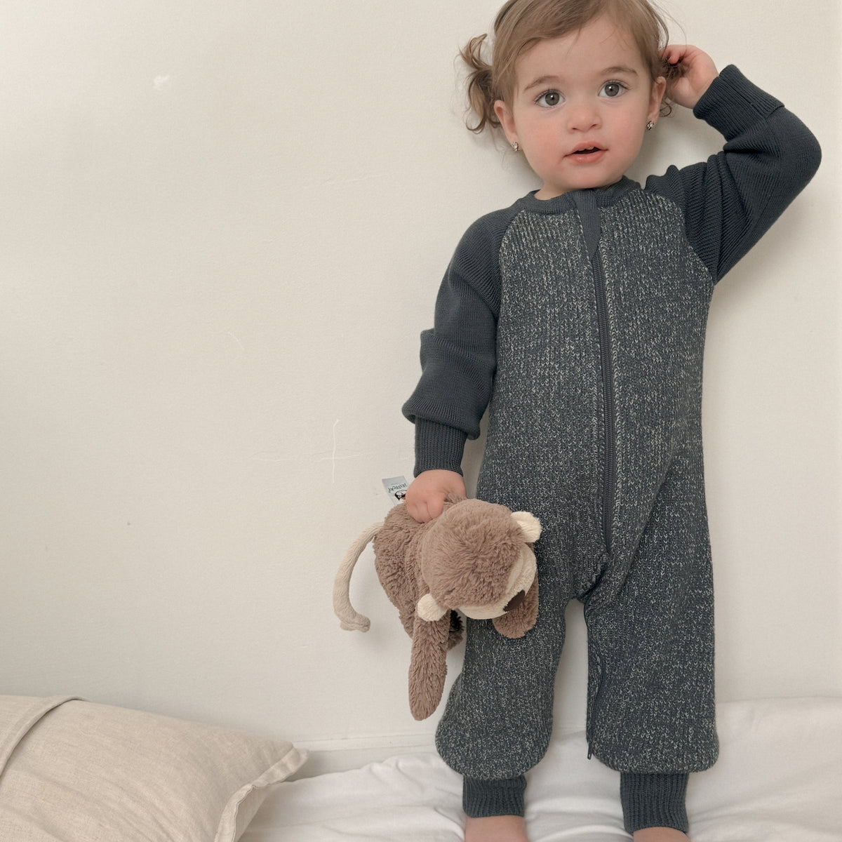 A toddler stands on a white bed wearing a dark gray and black long-sleeve one-piece romper, holding a brown plush monkey toy against a plain off-white wall.